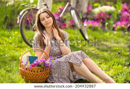 Beautiful young woman in dress sitting on grass with basket of flowers and reading book near old vintage bicycle