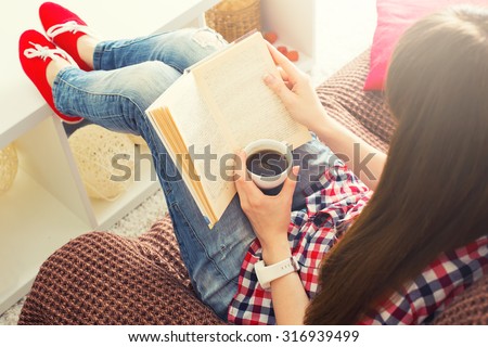 Woman sitting on the sofa reading a book holding her coffee mug in living room at home