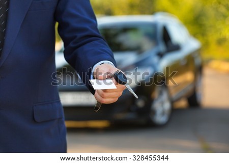 New car and business theme: a man in a black suit holds the keys of a new car and business card on a background of green tree