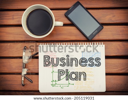 Notebook with text Business Plan inside on table with coffee, mobile phone and glasses.