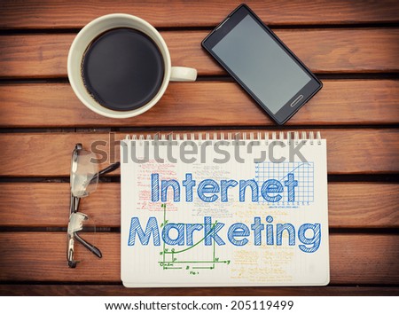 Notebook with text inside Internet Marketing on table with coffee, mobile phone and glasses.