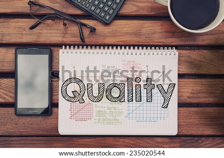 Notebook with text inside Quality on table with coffee, mobile phone and glasses.
