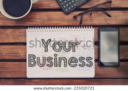 Notebook with text inside Your Business on table with coffee, mobile phone and glasses.