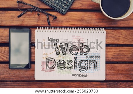 Notebook with text inside Web Design on table with coffee, mobile phone and glasses.