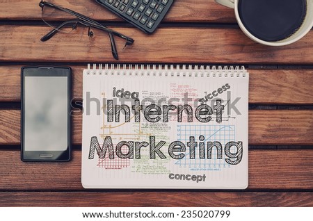 Notebook with text inside Internet Marketing on table with coffee, mobile phone and glasses.