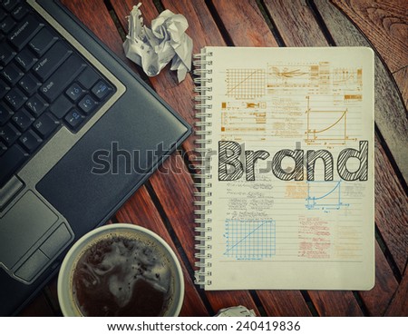 Notebook with text inside Brand on table with coffee, laptop pc and crumpled sheets