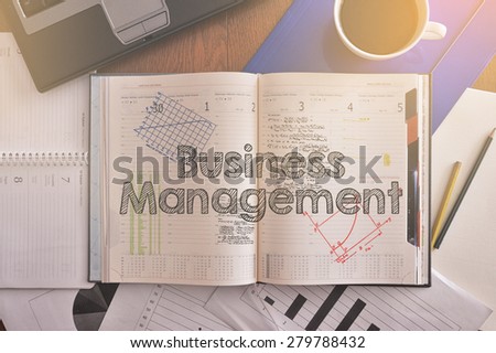 Notebook with text inside Business Management on table with coffee, some diagrams on paper and laptop