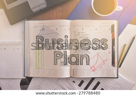 Notebook with text inside Business Plan on table with coffee, some diagrams on paper and laptop