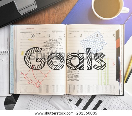 Notebook with text inside Goals on table with coffee, laptop and some sheet of papers with charts and diagrams