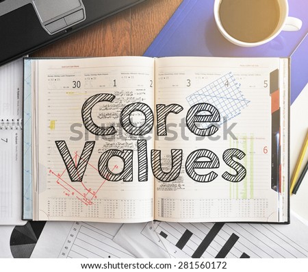 Notebook with text inside Core Values on table with coffee, laptop and some sheet of papers with charts and diagrams