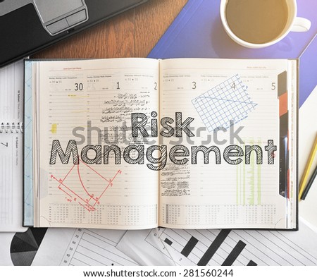 Notebook with text inside Risk Management on table with coffee, laptop and some sheet of papers with charts and diagrams
