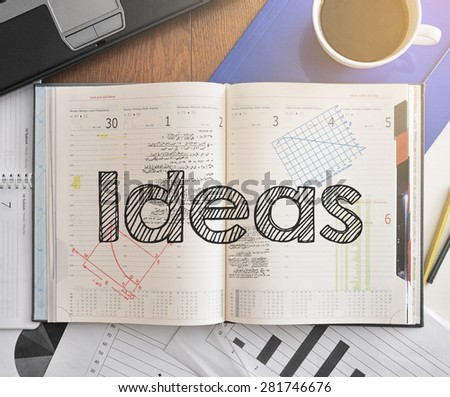 Notebook with text inside Ideas on table with coffee, some diagrams on paper and laptop