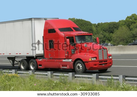 Red and white truck rolling down the highway with blue sky above and green grass below.