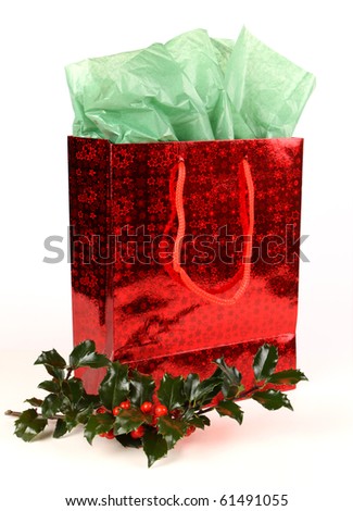 Red shiny Christmas gift bag with green tissue and a sprig of holly with red berries on white with soft shadow.