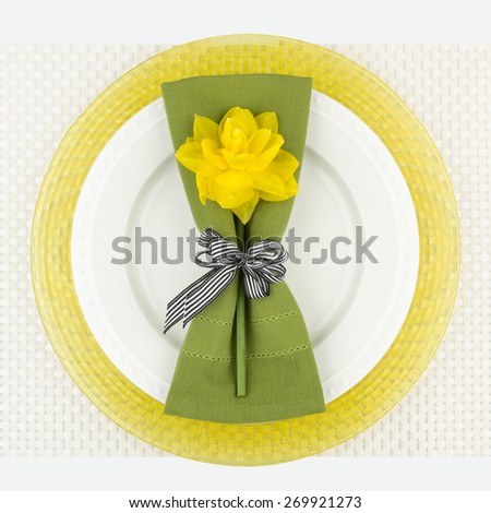 Simple place setting arrangement with yellow daffodil flower, green napkin, white plate and yellow charger on woven placemat. Close-up bird\'s eye view.