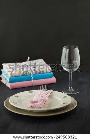Table setting: plate, fork, knife, pink napkin, glass with stack of books on dark wooden table