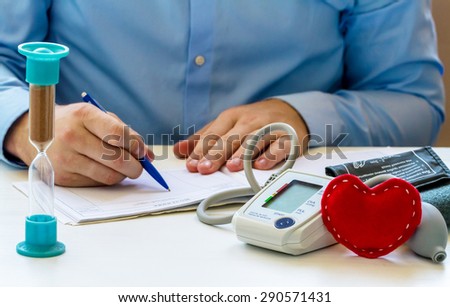 Doctor sitting at the white table with digital blood pressure monitor, red soft heart, hourglass  and writing a prescription