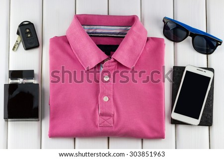 Top view of gentlemanly set: pink T-shirt, perfume, sunglasses, car keys, passport and smartphone on white wooden background