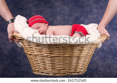 Newborn baby boy sleeping on a white blanket in a wicker basket. The basket is being held by his parents. Infant is wearing a red knit cap and diaper cover. Soft focus with a shallow depth of field.