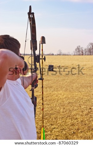 Young man in a white t-shirt aiming loaded bow at target in field