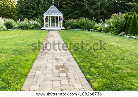 A white wedding gazebo on green lawn at the end of a stone pathway