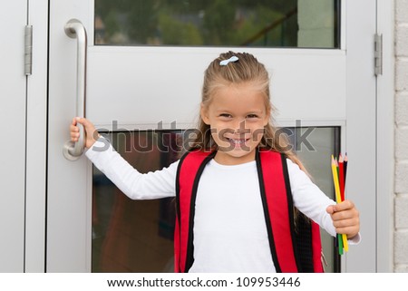 A Little Preteen Schoolgirl Holding a Handle of a School Door Showing her Pencils