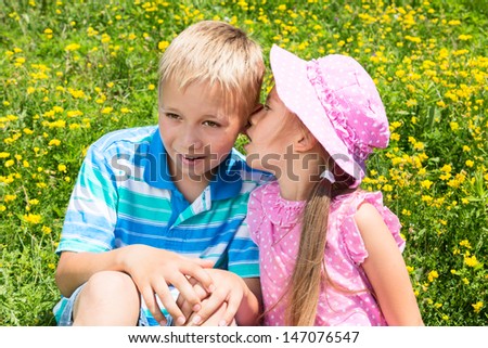 Two cute kids sitting on a grass with wildflowers, the girl is trying to whisper something to the boy\'s ear