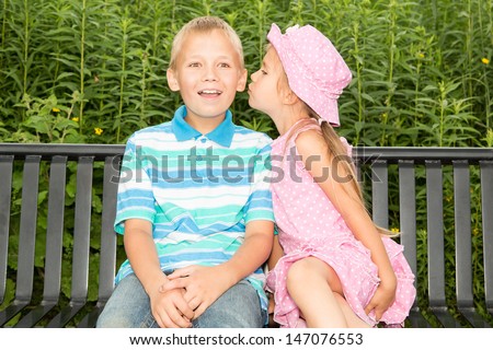 Two cute kids sitting on a park bench, the girl is trying to whisper something to the boy's ear, or maybe even to kiss him in the cheek !