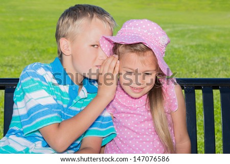 Two cute kids sitting on a park bench, the boy is trying to whisper something to the girl's ear looking at the camera with one eye at the same time
