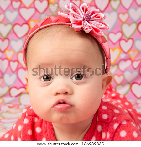 An adorable baby girl wearing a red headband with a decorative flower, lying on a blanket with hearts design print