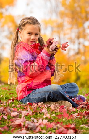 Beautiful young girl sitting on a grass with colorful autumn leaves, holding some red maple leaves in her hands