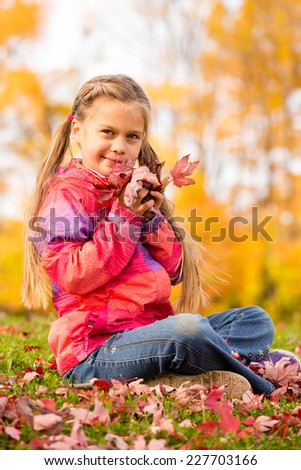 Beautiful young girl sitting on a grass with colorful autumn leaves, holding some red maple leaves close to her face