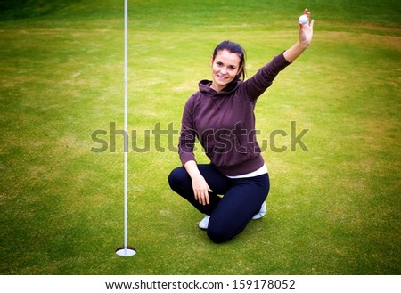 Smiling young woman golf player on green holding ball giving Thumbs Up sign