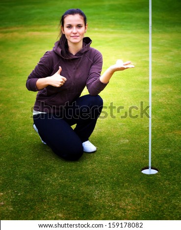 Smiling young woman golf player on green holding ball giving Thumbs Up sign