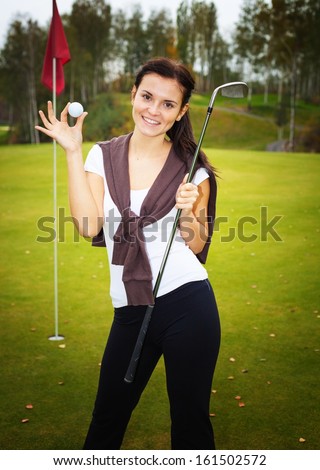 Smiling young woman golf player on green with ball and club near cup flag