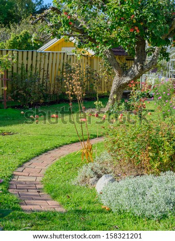 Autumn garden with brick path, flowers and fallen apples on the grass.