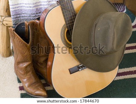 Brown cowboy boots and hat hanging off the neck of a guitar which is leaning against a colorful blanket.