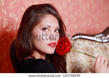 Beautiful girl in black dress holds red rose close to face and looks at camera.