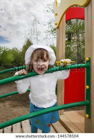 Cute little girl wearing white blouse stands on playground and looks at camera