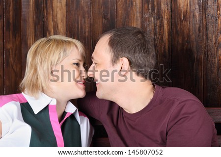 Happy wife and husband touch each other noses and smile near wooden wall.