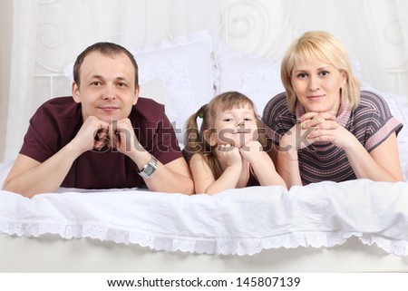Happy father, little daughter and mother lie on white double bed and prop up chin by hands. Focus on woman.