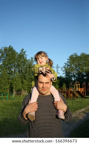 Little daughter sits on shoulders of father and keeps his head outdoors.