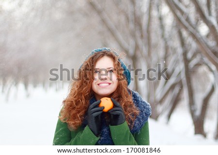 Beautiful girl in blue beret holds mandarin and looks up at winter day in park