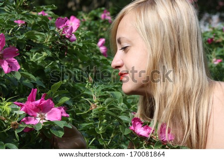 Beautiful blonde girl looks at pink flowers on bush in park at summer day