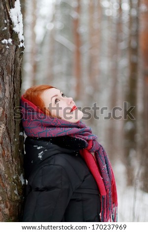 Smiling girl stands next to tree and looks up outdoor at winter day in forest