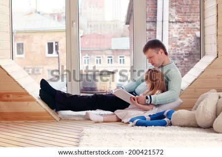 Pretty little blonde girl and father sit near window and read book at home