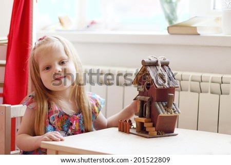 Pretty little girl sits at table with small wooden toy house and smiles