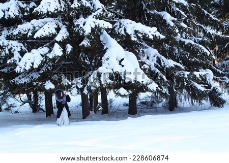 Beautiful girl in fur coat hides under big snowy spruces in winter forest