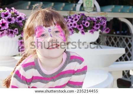 Happy little girl with pictured purple butterfly on face near pots of flowers outdoor