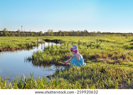 Beautiful cute little girl in blue dress sitting by creek with stick in hand
